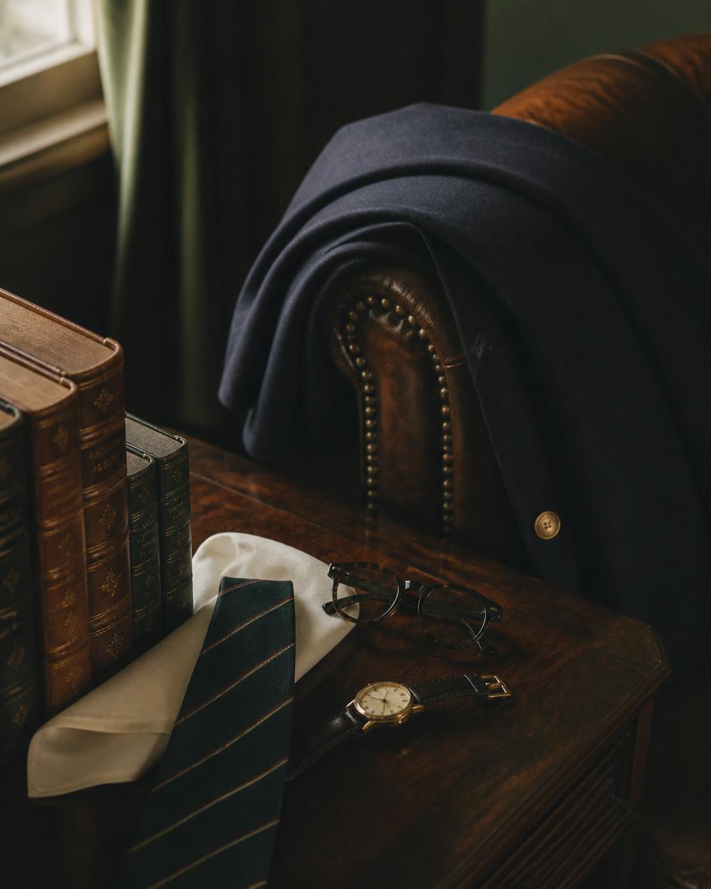 A navy blazer draped over a leather club chair beside leather-bound books, a silk tie and a vintage watch